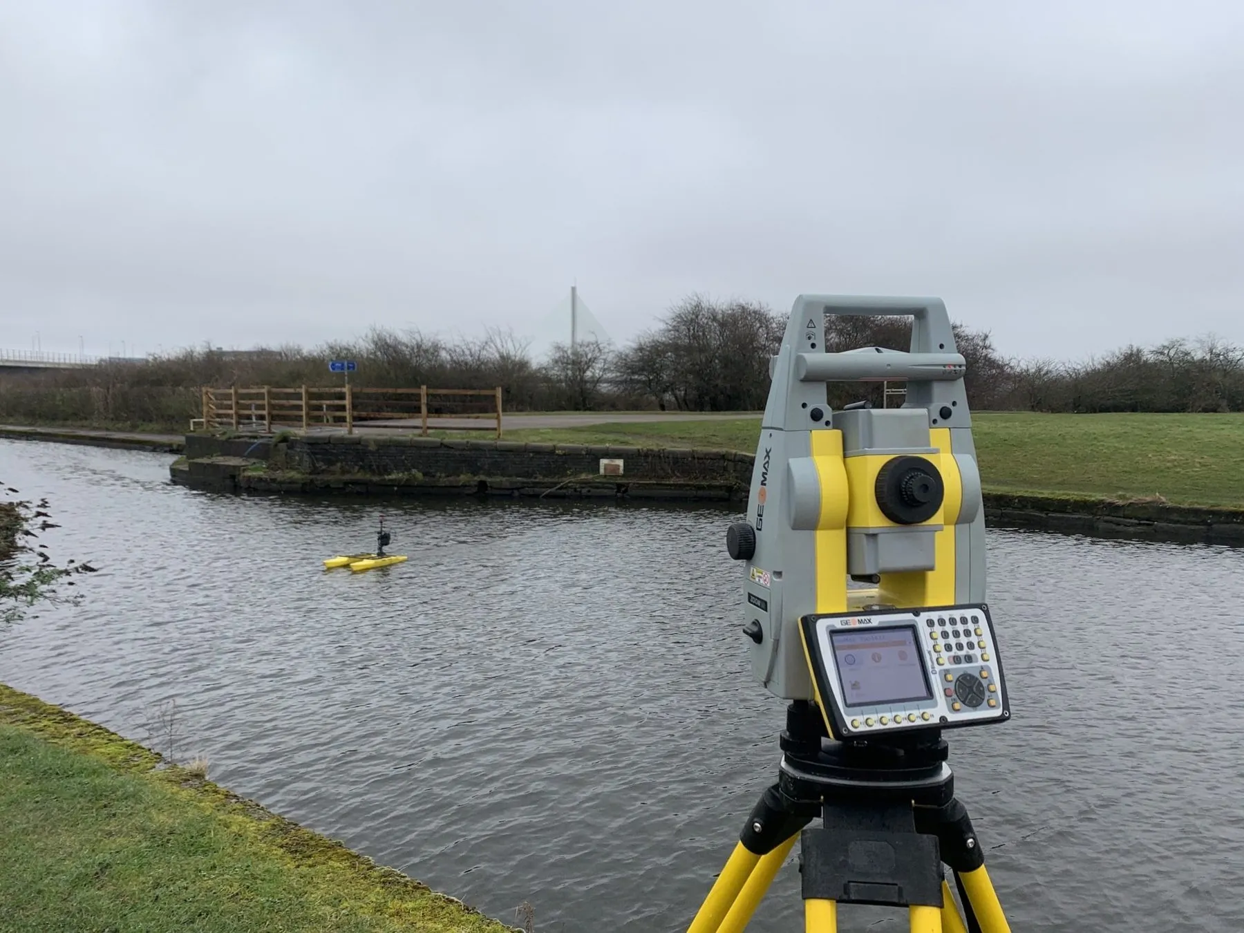 Total station on a riverbank tracking a remote survey boat during a bathymetric survey in Cambridge